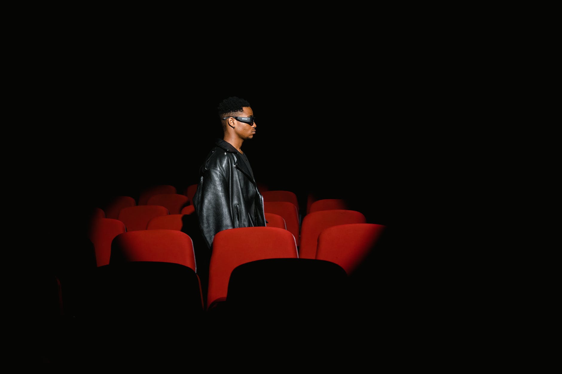 man black leather jacket standing behind red theatre seat