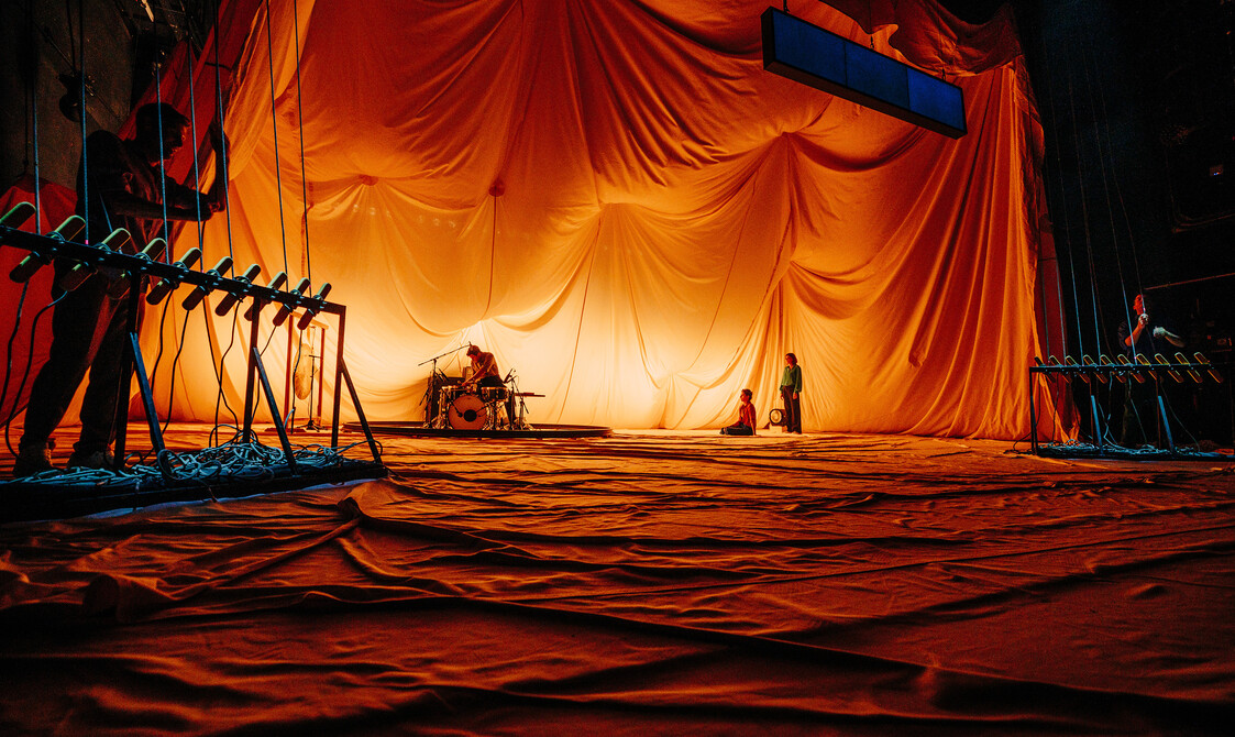 Um baterista senta-se diante de seu kit enquanto alguém está sentado no chão e outro está de pé, olhando para eles.  O pano de fundo é uma grande tenda de lona que se acomoda em ondas dramáticas acima deles, iluminada com um brilho suave e quente que deixa o resto da tenda na sombra.