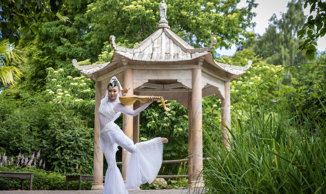 Uma dançarina chinesa segura seu instrumento e dança em um traje branco com calças largas em frente à rotunda chinesa no Jardim Botânico de Edimburgo