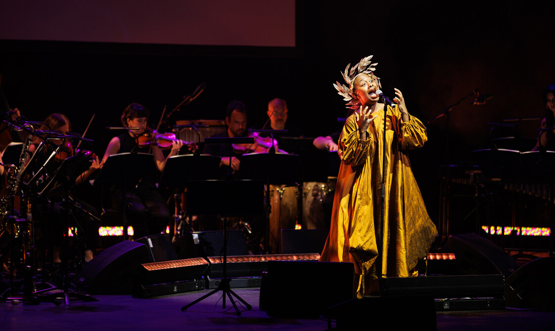 Uma mulher canta em frente a um microfone usando um grande vestido dourado e um adorno de cabeça dourado.  Atrás dela está uma banda tocando instrumentos.