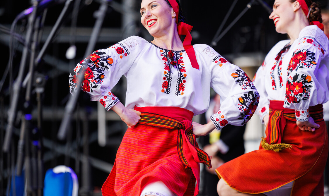 Duas mulheres dançam e sorriem no palco, com os joelhos levantados e as mãos na cintura.  Elas usam trajes tradicionais, com blusas bordadas e saias vermelhas.